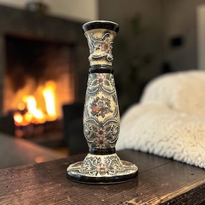 May include: A ceramic candlestick holder featuring a cream-colored base with a crackle glaze finish. The surface is decorated with intricate black, pink, and orange floral patterns and scrolling leaf motifs, accented by solid black bands at the top and base.