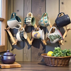 May include: A wooden mug rack displays eight ceramic mugs with various designs and colors. A purple teapot sits on a wooden board, and a wicker basket holds a lemon and herbs. The mugs are hanging on a wooden rack.