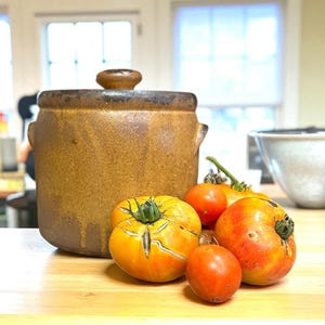 May include: A brown ceramic crock with a matching lid and side handles sits next to a cluster of fresh tomatoes. The tomatoes vary in size and color, ranging from bright red to yellow-orange, with some showing natural skin cracks and green stems.