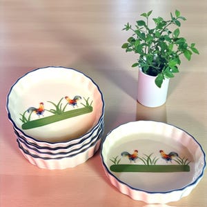May include: A set of white ceramic tart dishes with a blue scalloped edge. Each dish features a colorful rooster design with green grass. A small white vase with green leaves is also present.