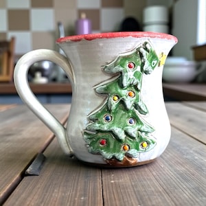 May include: A ceramic mug with a white base, a red rim, and a curved handle. The mug features a raised green Christmas tree design with colorful ornaments. The mug is sitting on a wooden surface.