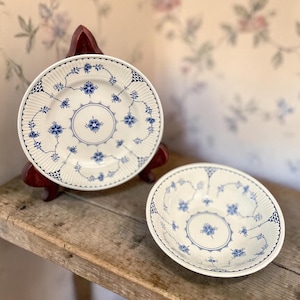 May include: Two white ceramic plates with blue floral designs. One plate is on a wooden stand. The plates have a scalloped edge and a central floral motif. The background is a floral wallpaper.