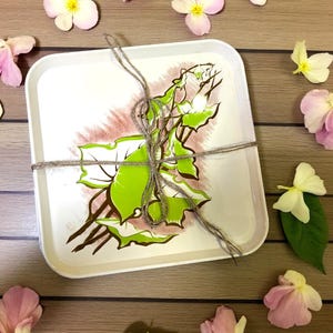 May include: A white rectangular tray with a green and brown floral design, tied with twine. Scattered around the tray are pink and white flowers and green leaves, all on a wooden surface.
