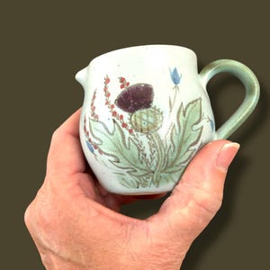 May include: A small, off-white ceramic creamer with a thistle design. The creamer has a spout and a curved handle. The thistle design features purple, green, red, and blue accents. The creamer is held in a hand.