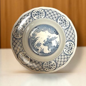 May include: A white and blue ceramic plate with a scalloped edge. The plate features a central design of a bird in a landscape, surrounded by floral and geometric patterns. The plate is approximately 10 inches in diameter.