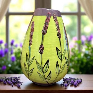 May include: A decorative, teardrop-shaped vase with a light green body and a purple rim. The vase features hand-painted lavender sprigs with black stems and green leaves. The vase is displayed on a wooden surface.