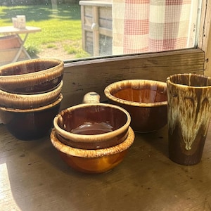 May include: A collection of vintage brown and tan ceramic bowls and a tall tumbler. The bowls are stacked and arranged on a wooden surface. The tumbler has a dripped glaze design. The bowls have a handle.