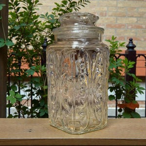 May include: A clear, rectangular glass jar with a lid, featuring an embossed floral design. The jar is sitting on a wooden surface, with greenery and a brick wall in the background. The jar is approximately 8 inches tall.