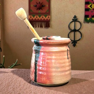 May include: A handmade ceramic jar with a wooden spoon. The jar is cylindrical with a lid and features a color gradient of red, pink, and beige. A black vertical stripe and a wooden spoon add contrast.