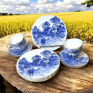 May include: A set of blue and white porcelain teacups and saucers with a country cottage scene. The set is displayed on a wooden table with a field of wheat in the background.