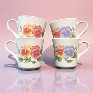 May include: Four white ceramic mugs with curved handles are stacked in pairs. Each mug features a painted floral design with yellow, pink, and purple pansies and green leaves. The mugs are set against a pink background.