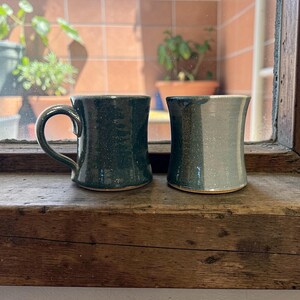 May include: Two handmade ceramic mugs. One is dark green with a handle, the other is a two-tone gray and green. Both mugs have a speckled texture and sit on a wooden surface near a window with plants visible in the background.
