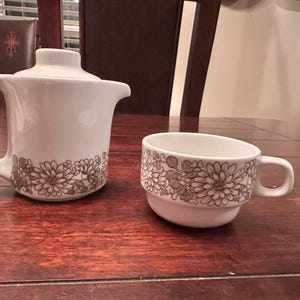 May include: White ceramic teapot with lid and matching teacup. Both pieces feature a brown floral pattern around the base. The teapot has a handle and spout. The teacup has a handle and a stacked base.