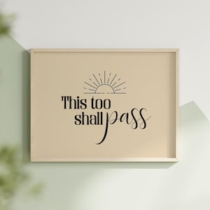 May include: Framed print with the inspirational quote "This too shall pass" in black script and a sunburst design. The artwork is set against a beige background, framed in a light wood, and displayed on a white and green wall.