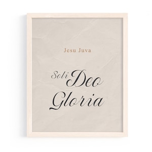 May include: Framed print with the text "Jesu Juva" in a light brown font above "Soli Deo Gloria" in a black script font. The background is a textured beige color, and the frame is a light wood color.