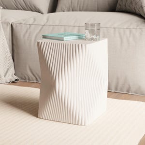 May include: A white, sculptural side table with a textured, wave-like design. A glass of water and a small book rest on top. The table is placed on a cream-colored rug in front of a beige sofa.