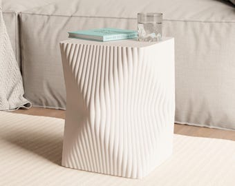 May include: A white, sculptural side table with a textured, wave-like design. A glass of water and a small book rest on top. The table is placed on a cream-colored rug in front of a beige sofa.