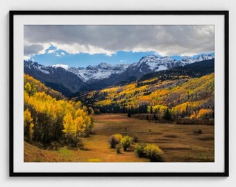 Puede incluir: Fotografía enmarcada de un paisaje de valle de montaña. Los árboles de color amarillo dorado bordean el fondo del valle y las laderas, con montañas cubiertas de nieve en el fondo bajo un cielo nublado. La fotografía está en un marco negro.