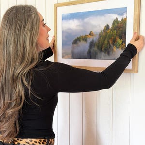 Puede incluir: Una fotograf&iacute;a enmarcada de un paisaje monta&ntilde;oso con &aacute;rboles y niebla. La fotograf&iacute;a est&aacute; en un marco de madera clara con un passe-partout blanco. La imagen se sostiene contra una pared blanca.