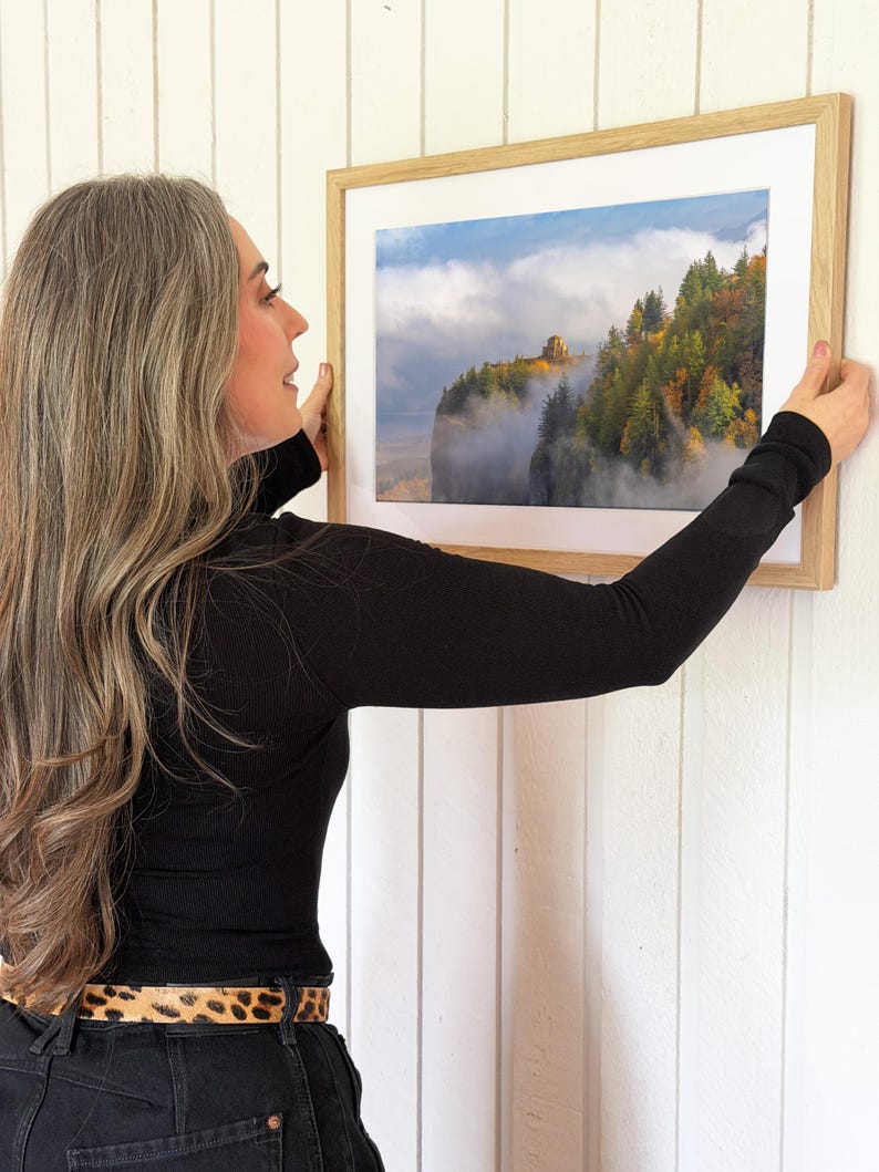 Puede incluir: Una fotograf&iacute;a enmarcada de un paisaje monta&ntilde;oso con &aacute;rboles y niebla. La fotograf&iacute;a est&aacute; en un marco de madera clara con un passe-partout blanco. La imagen se sostiene contra una pared blanca.