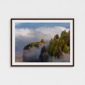 Puede incluir: Impresi&oacute;n enmarcada de un paisaje con un acantilado y una estructura de piedra en la cima de una cresta arbolada. La escena est&aacute; envuelta en niebla, con un cielo azul visible. Los &aacute;rboles muestran colores oto&ntilde;ales.