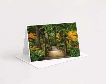 Puede incluir: Una tarjeta de felicitación plegada con una fotografía de un puente de madera arqueado que cruza un sendero en un bosque frondoso. La escena incluye árboles altos, follaje verde vibrante y parches de hojas amarillas y naranjas.