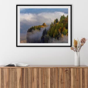 Puede incluir: Fotograf&iacute;a enmarcada de un paisaje de acantilado con una estructura de piedra en la cima, rodeada de &aacute;rboles con colores oto&ntilde;ales. La escena est&aacute; parcialmente oscurecida por la niebla, con un cielo azul arriba. La fotograf&iacute;a se exhibe en un mueble de madera.