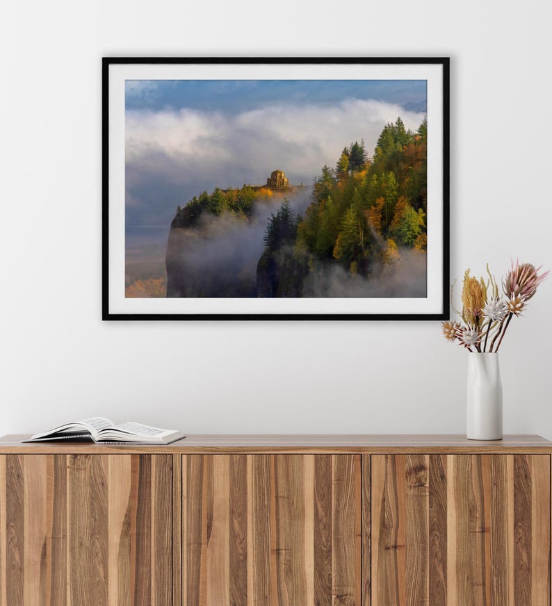 Puede incluir: Fotograf&iacute;a enmarcada de un paisaje de acantilado con una estructura de piedra en la cima, rodeada de &aacute;rboles con colores oto&ntilde;ales. La escena est&aacute; parcialmente oscurecida por la niebla, con un cielo azul arriba. La fotograf&iacute;a se exhibe en un mueble de madera.