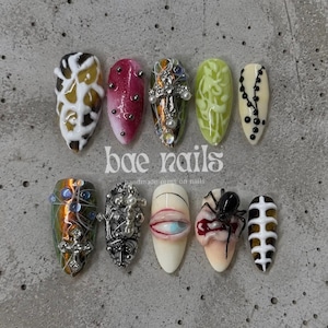 May include: A set of ten press-on nails featuring diverse designs including a brown and white marbled pattern, a pink gradient with silver studs, a cross charm, green vine art, black dots, a spider, a realistic eye, and skeletal rib motifs.