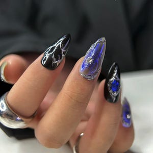 Peut inclure: Main montrant de longs faux ongles pointus. Les ongles sont noirs, iris&eacute;s et bleus, avec des accents argent&eacute;s et des pierres pr&eacute;cieuses. Le texte "bae nails" et "handmade press on nails" est visible en arri&egrave;re-plan.