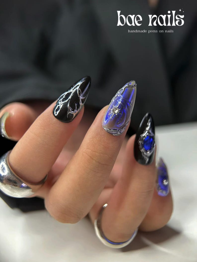 Peut inclure: Main montrant de longs faux ongles pointus. Les ongles sont noirs, iris&eacute;s et bleus, avec des accents argent&eacute;s et des pierres pr&eacute;cieuses. Le texte "bae nails" et "handmade press on nails" est visible en arri&egrave;re-plan.