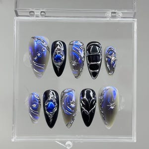Peut inclure: Un ensemble de dix faux ongles dans un &eacute;tui en plastique transparent. Les ongles pr&eacute;sentent divers motifs, notamment des accents argent&eacute;s, des tourbillons bleus et argent&eacute;s et des bases noires et bleues. Les ongles sont de forme ovale.