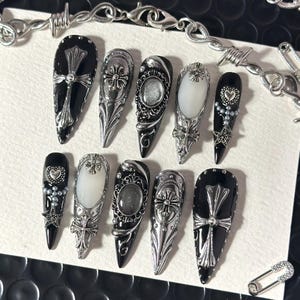 Puede incluir: Un juego de diez uñas postizas en forma de estilete con una paleta de colores negro y plata. Los diseños incluyen emblemas de cruces metálicas, marcos de filigrana plateada, acentos ovalados blancos, corazones y estrellas.