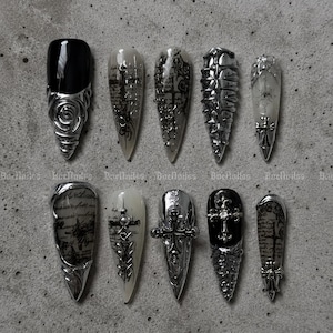 Pode incluir: Um conjunto de dez unhas postiças em formato stiletto com uma paleta de cores monocromática de preto, branco e prata metálico. Os designs incluem detalhes metálicos prateados em 3D, motivos de cruz, rosas e escrita estilo vintage.