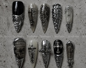 Könnte beinhalten: Ein Set aus zehn stilettoförmigen Press-on-Nägeln in einer monochromen Farbpalette aus Schwarz, Weiß und Metallic-Silber. Die Designs umfassen detaillierte 3D-Metallakzente in Silber, Kreuzmotive, Rosenverzierungen und eine Vintage-Schrift.