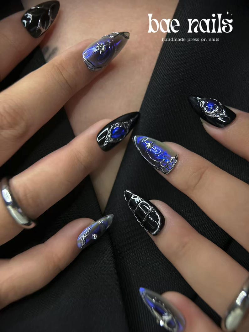 Peut inclure: Gros plan sur plusieurs faux ongles longs et pointus. Les ongles sont noirs avec des accents argent&eacute;s et bleus. Certains ongles ont un effet bleu iris&eacute;. Le texte "bae nails handmade press on nails" est visible en haut &agrave; droite.