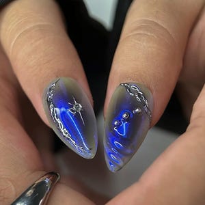 Peut inclure: Gros plan sur deux faux ongles en forme d'amande avec un motif bleu et argent&eacute;. Les ongles pr&eacute;sentent un effet d&eacute;grad&eacute;, passant d'une base transparente &agrave; une teinte bleu profond. Des accents argent&eacute;s en forme de cha&icirc;ne et des ornements en forme d'&eacute;toile ajoutent des d&eacute;tails. Le texte "bae nails handmade press on nails" est visible.