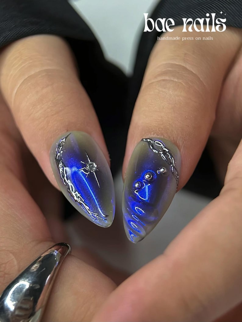 Peut inclure: Gros plan sur deux faux ongles en forme d'amande avec un motif bleu et argent&eacute;. Les ongles pr&eacute;sentent un effet d&eacute;grad&eacute;, passant d'une base transparente &agrave; une teinte bleu profond. Des accents argent&eacute;s en forme de cha&icirc;ne et des ornements en forme d'&eacute;toile ajoutent des d&eacute;tails. Le texte "bae nails handmade press on nails" est visible.