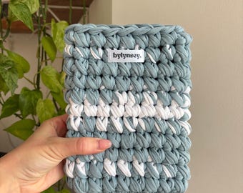 May include: A hand-crocheted rectangular basket in shades of light blue and white. The basket features a textured stitch pattern with a horizontal white stripe. The label "bylinsey." is visible on the front. The basket is held by a hand.