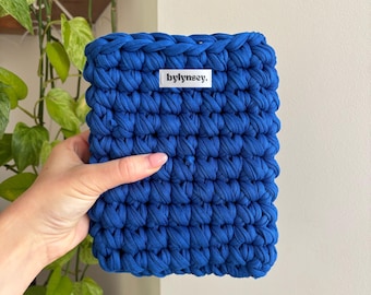 May include: A vibrant blue, hand-crocheted pouch. The rectangular bag features a chunky knit design and a small white label with the text "bylinsey." The bag is held in a person's hand, with green plants in the background.