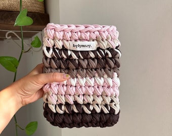 Puede incluir: Un estuche rectangular de ganchillo hecho de hilo de tela grueso con rayas horizontales en rosa claro, beige, blanco y marrón oscuro. Una pequeña etiqueta de tela blanca con el texto negro bylynsey. está cosida en la parte superior central.