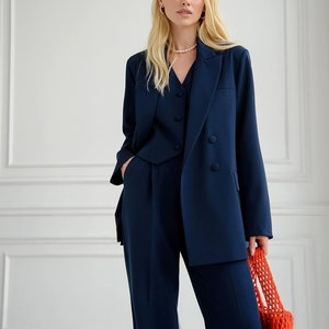 May include: A woman wearing a navy blue blazer and matching pants. She is wearing a pearl necklace and has a red crocheted bag in her hand.