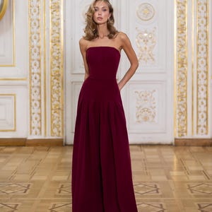 May include: A floor-length, strapless jumpsuit in a deep burgundy color. The garment features a fitted bodice and wide-leg trousers with subtle pleats at the waist, creating a clean, structured silhouette that extends to the floor.