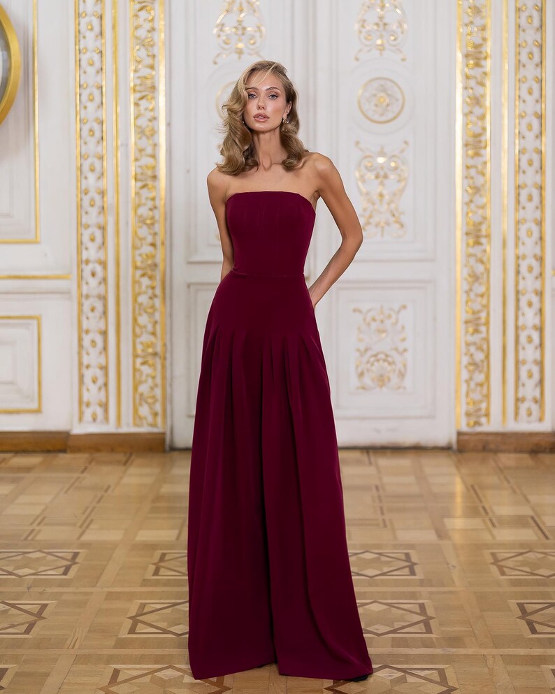 May include: A floor-length, strapless jumpsuit in a deep burgundy color. The garment features a fitted bodice and wide-leg trousers with subtle pleats at the waist, creating a clean, structured silhouette that extends to the floor.