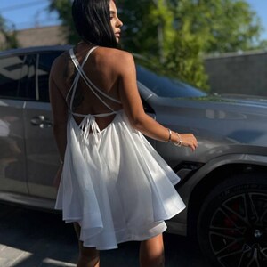 Puede incluir: Vestido blanco con espalda descubierta, tirantes finos y falda acampanada. El vestido es de tejido ligero y tiene un cierre de lazo en la espalda. La persona lleva sandalias de plataforma negras.