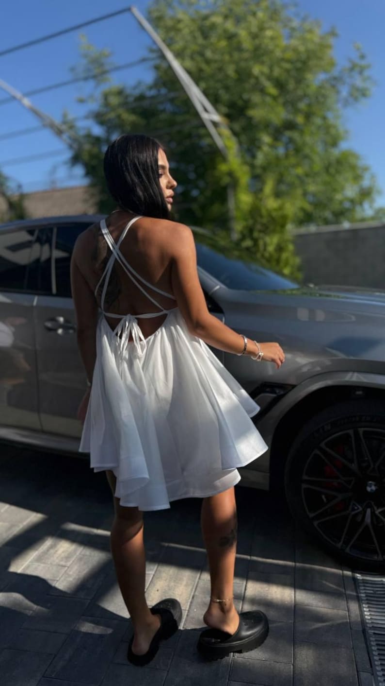 Puede incluir: Vestido blanco con espalda descubierta, tirantes finos y falda acampanada. El vestido es de tejido ligero y tiene un cierre de lazo en la espalda. La persona lleva sandalias de plataforma negras.