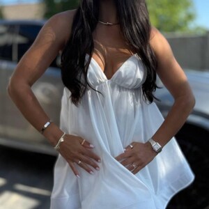 Puede incluir: Un vestido blanco y fluido con tirantes finos. El vestido tiene un escote en V profundo y un dobladillo corto. La persona lleva un collar de oro, un reloj y varios anillos y pulseras. El fondo incluye un coche y un cielo soleado.