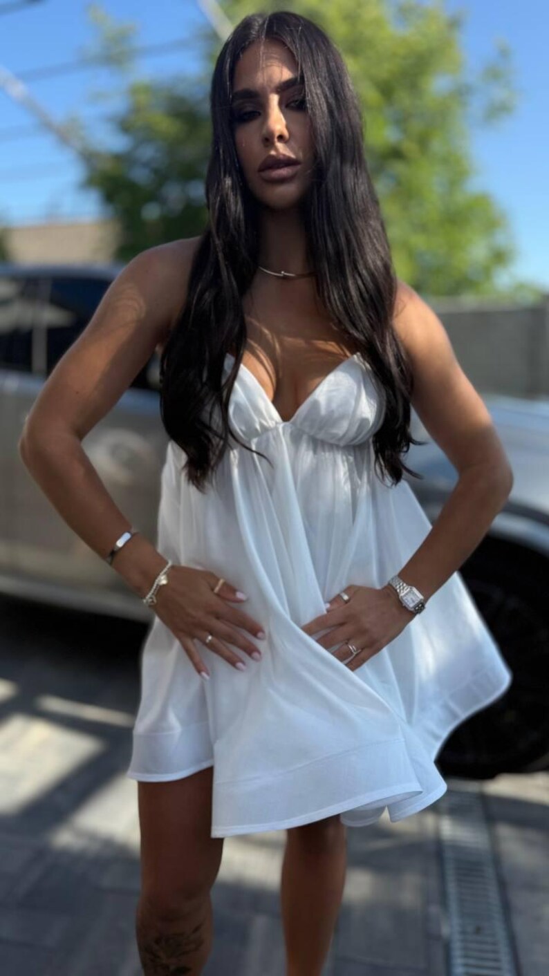 Puede incluir: Un vestido blanco y fluido con tirantes finos. El vestido tiene un escote en V profundo y un dobladillo corto. La persona lleva un collar de oro, un reloj y varios anillos y pulseras. El fondo incluye un coche y un cielo soleado.