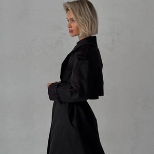 May include: A black trench coat with a red stitch detail. The coat has a long, flowing silhouette and a classic collar.