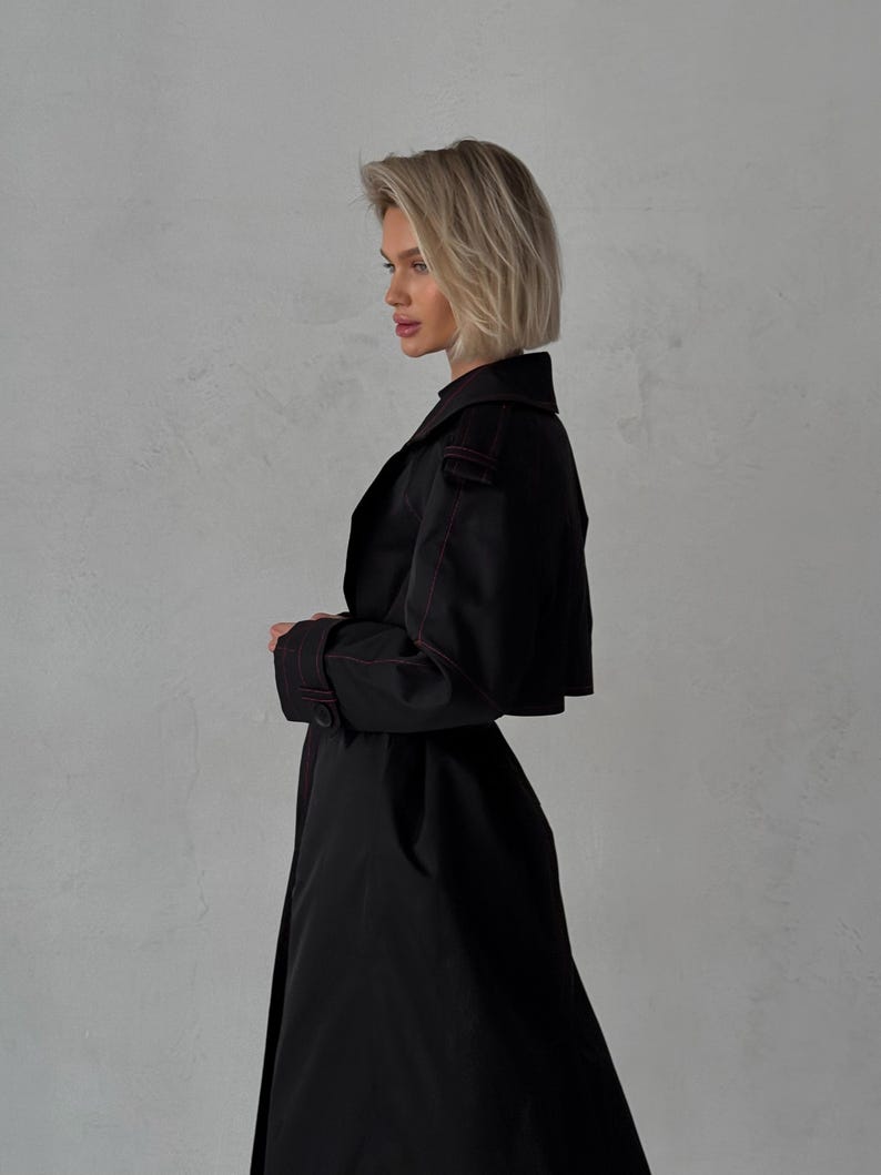 May include: A black trench coat with a red stitch detail. The coat has a long, flowing silhouette and a classic collar.
